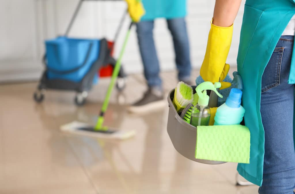 Middle-aged woman in green apron with cleaning supplies, laundry detergent, organized bucket, cleaning brush, mop with red bucket in background.