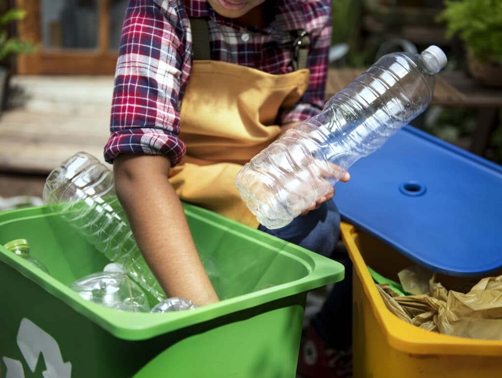 A dedicated SCS Group cleaner recycles plastic bottles, demonstrating her commitment to sustainability in a bustling commercial complex. A nearby trash can contrasts improper waste disposal. The image conveys shared responsibility for a clean and sustainable environment.
