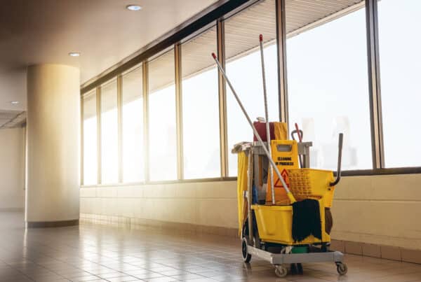 The image captures a bustling scene in a hallway where cleaning supplies are in action. At the center of the frame stands a yellow cart brimming with various cleaning products