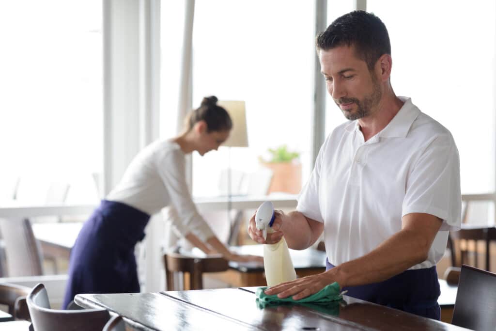 a restaurant where a pair of hospitality cleaners are cleaning a table. The man is wearing a white shirt, while the woman is wearing a white apron. The woman can be seen wiping the table with a cloth, while the man is looking at his phone.