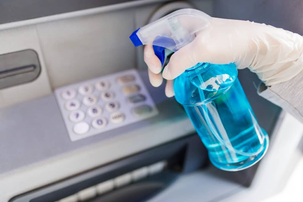 A bank cleaner in blue gloves sprays a blue liquid onto an ATM machine. This creates a feeling of mystery and urgency.