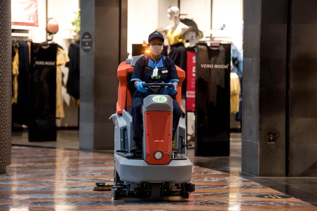A lively scene takes place in a busy shopping mall. A person rides a robot. Workers use cleaning machines. Fashion displays catch the eye.
