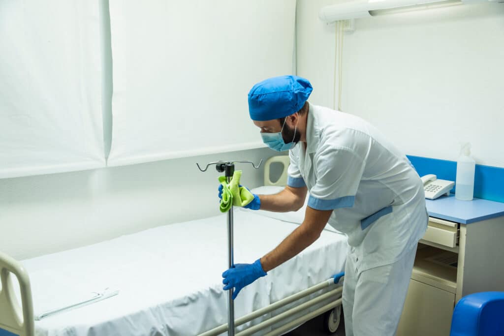 A cleaning staff member in a blue scrub hat carefully cleans a hospital bed. A nurse helps nearby, surrounded by important medical equipment and supplies.