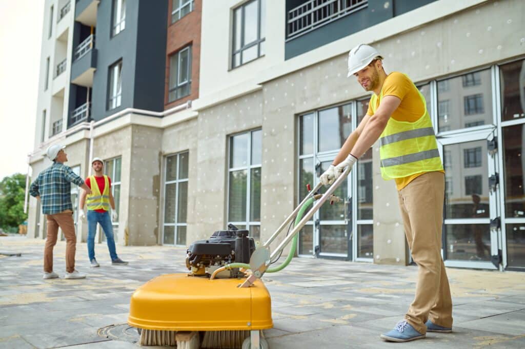 Construction workers sweep, grind, and excavate on a bustling site. Two workers stand by a building, one with a shovel and the other on his phone, while a group gathers in the background for a harmonious collaboration