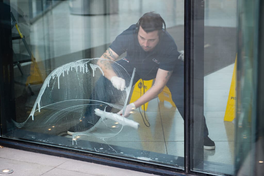 A Corporate cleaner wearing headphones meticulously cleans a glass door with a cloth, while a yellow sign stands on the sidewalk and text adds intrigue to the scene.