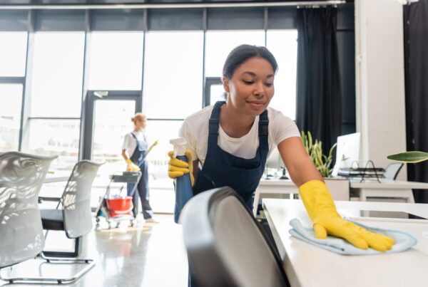 Smiling,Bi-racial,Woman,Cleaning,Office,Desk,With,Rag,Near,Colleague