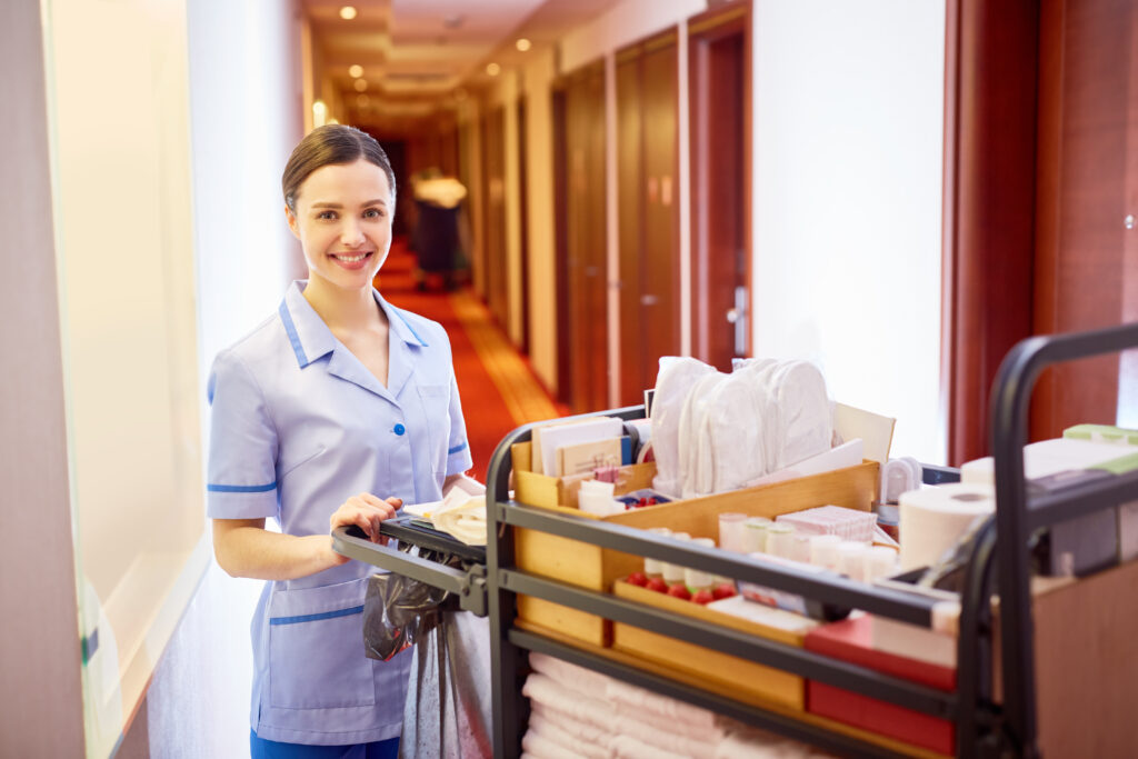 Young,Female Hotel Cleaner ,With,Tidy,Supplies,Standing,In,Hotel,Corridor