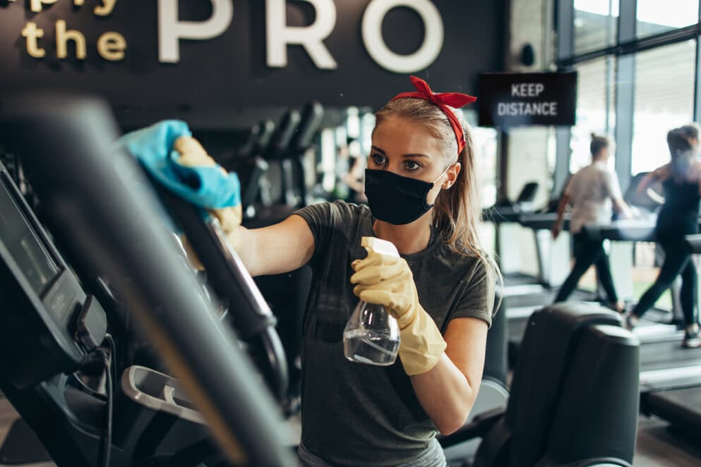 A woman wearing a mask and gloves carefully cleans a treadmill in a gym. She wants to keep the workout area safe.