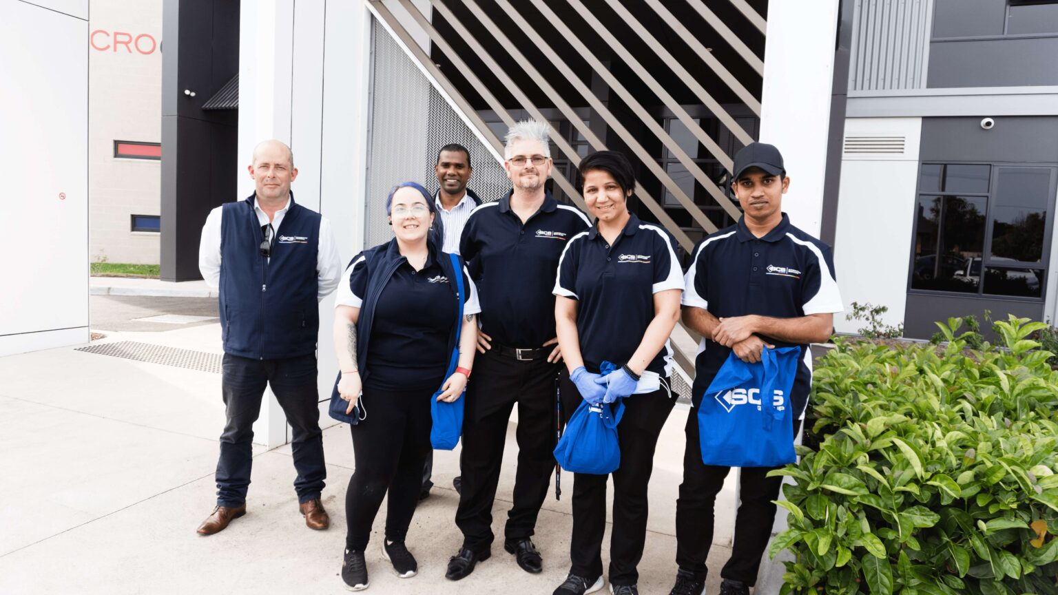 Five individuals in blue shirts and bags stand outside a building, representing the SCS group.