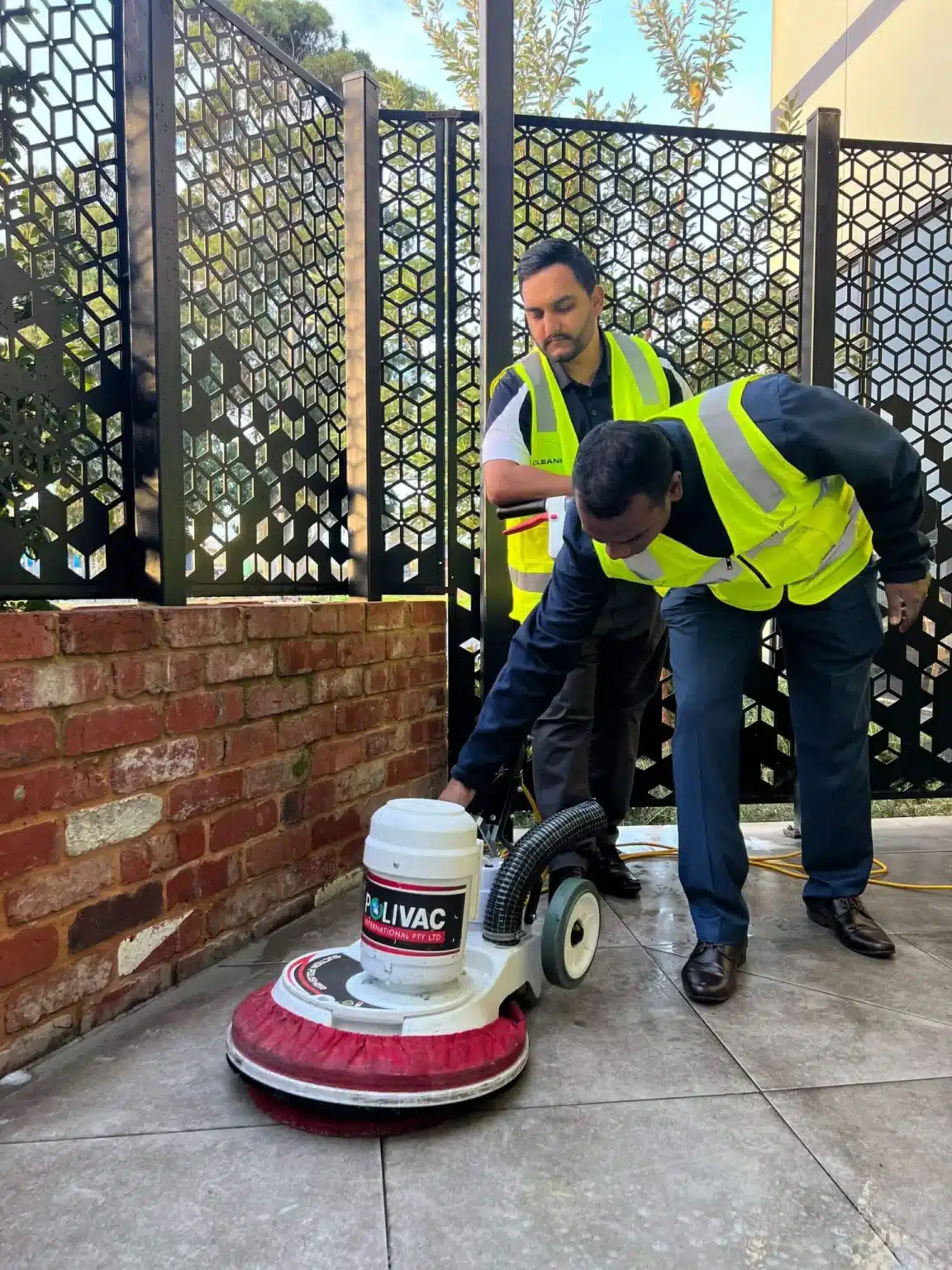 Two men in yellow vests are using a vacuum to clean a floor, demonstrating teamwork in maintaining cleanliness.