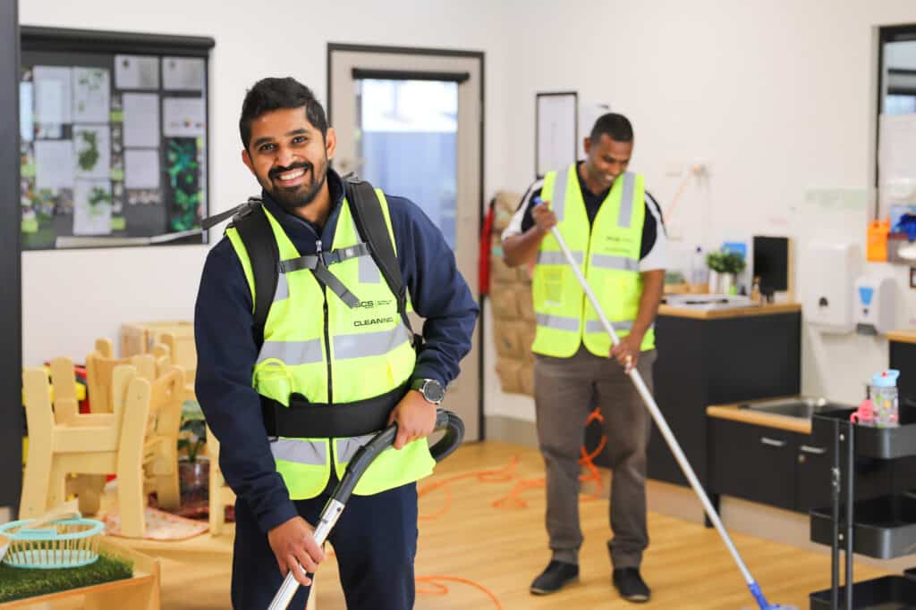 Two men in bright yellow vests are actively cleaning a room, contributing to a neat and well-maintained space.