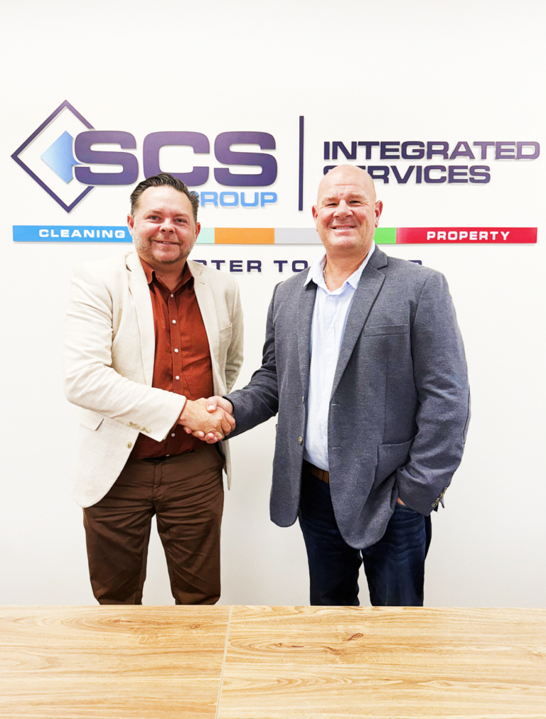 Two men in business attire shaking hands in front of an SCS Group Integrated Services wall sign.