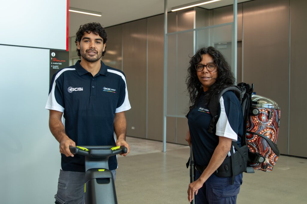 Two cleaners wearing GCS uniforms standing indoors with cleaning equipment, including a floor machine and a vacuum featuring an Indigenous design.