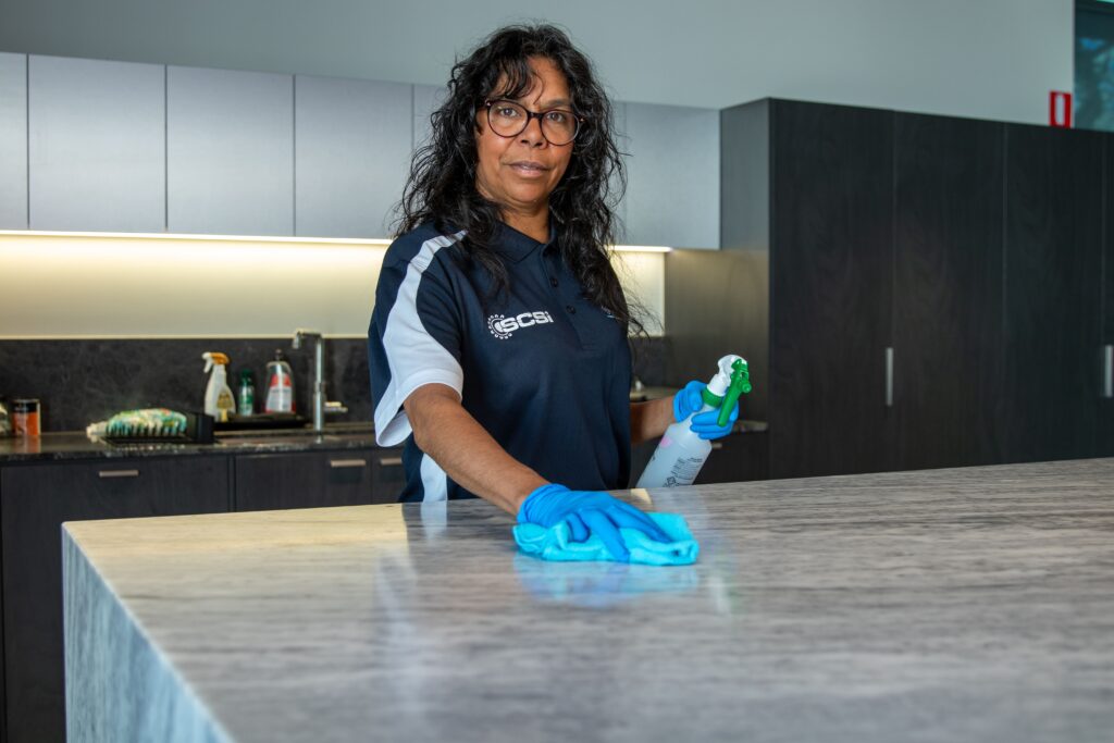 Cleaner wearing gloves and an SCS uniform wiping a marble countertop with a spray bottle in a modern kitchen setting.