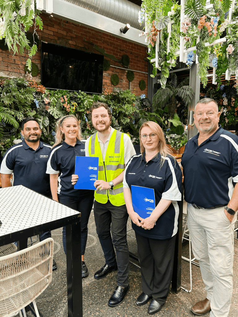 Team of five staff members standing together indoors with greenery behind them, holding blue SCS Cleaning Services folders and smiling.