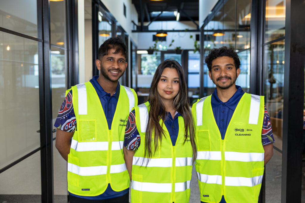 Three SCS Cleaning staff members in hi-visibility yellow safety vests and branded navy polo shirts smile at the camera while standing in the corridor of a modern commercial office building.