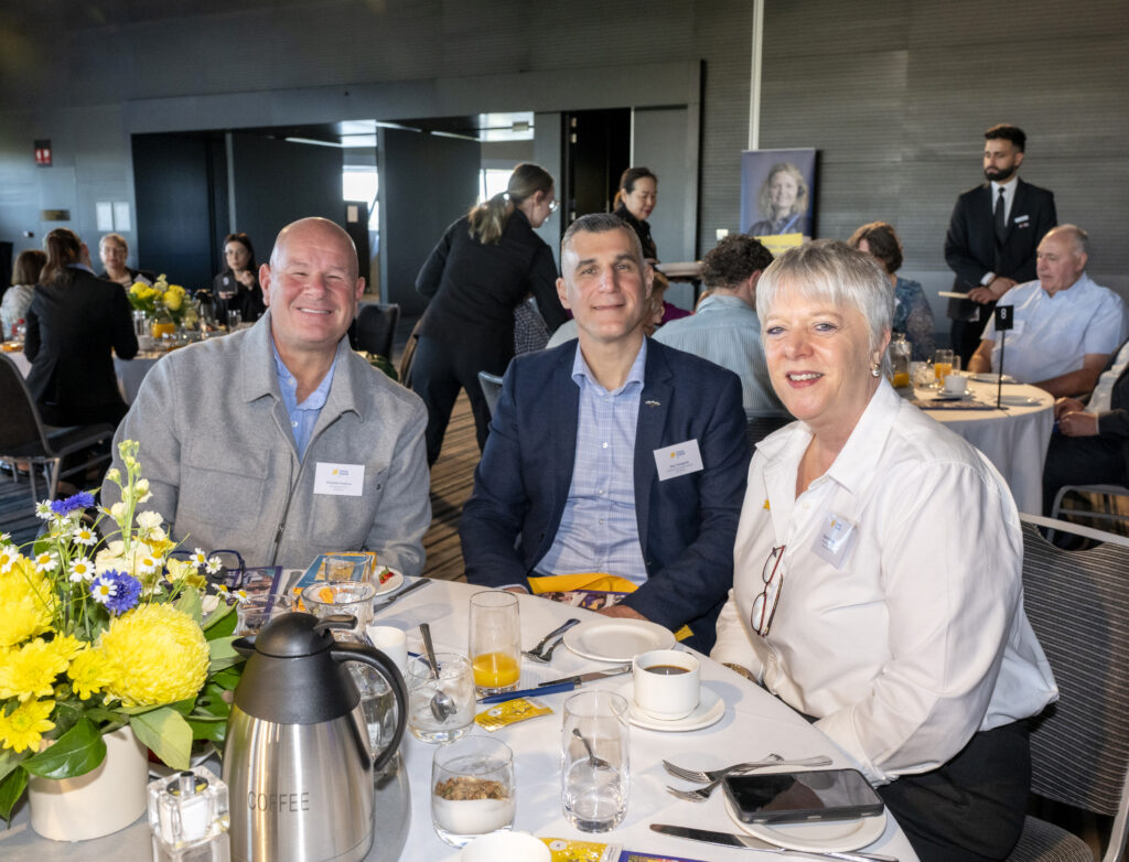 Three attendees at a Cancer Council breakfast event, seated at a table with yellow floral centre pieces
