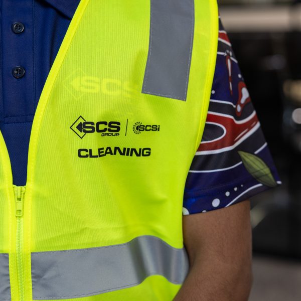 Close-up of a cleaning staff member wearing a hi-vis yellow safety vest with reflective strips over a navy polo shirt, branded with the SCS Group and SCSI logos above the word "Cleaning."